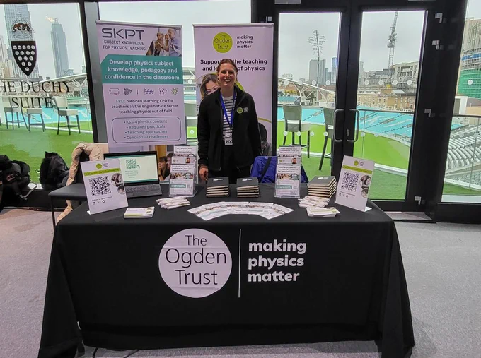 Ogden Trust at conference. A member of staff is still  with two banner stands behind her and table in front. The table is displaying leaflets and notebooks, and is covered with a black table cloth featuring the Ogden logo in white. There is a view of the Oval Cricket ground from the windows/doors behind the stand.