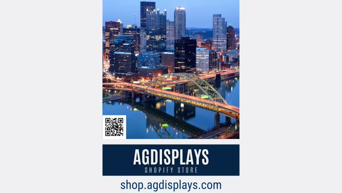 Evening shot of Pittsburgh's downtown skyline with lit-up high-rises and the yellow bridge over the calm river, complete with a QR code and branding for "AGDISPLAYS Shopify Store shop.agdisplays.com" in the foreground.