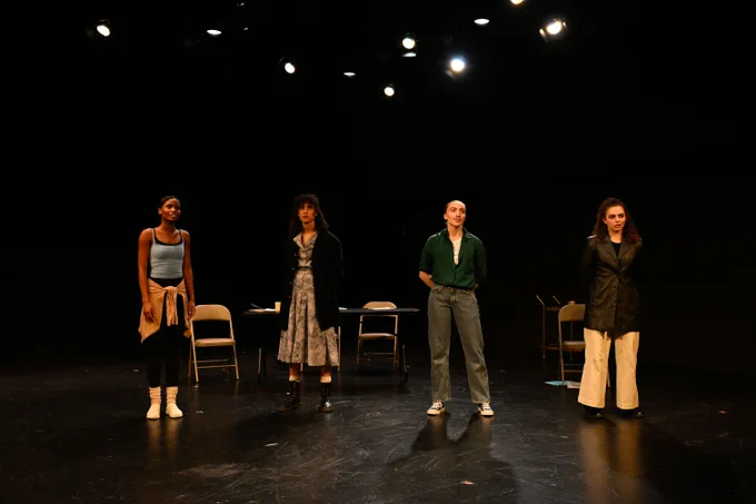 Four actors standing on stage in a theater setting, each dressed in distinct styles, facing the audience.