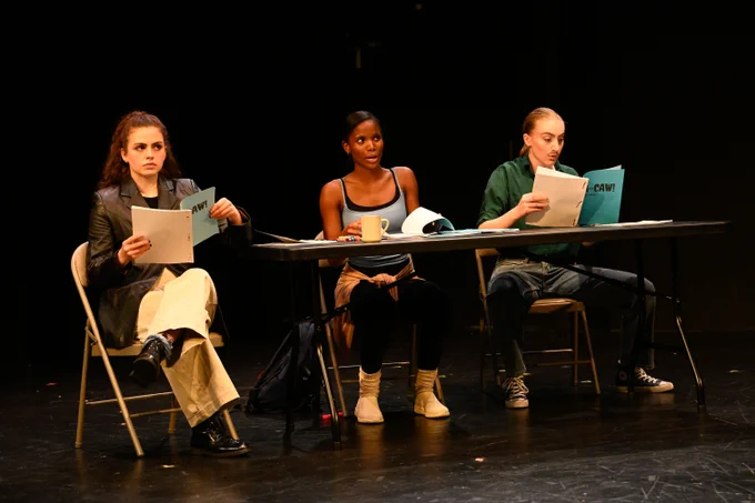 Three individuals are seated at a table during a script reading session in a theater setting. They each hold scripts and appear focused on their reading.