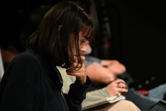 Person sitting in a dark room concentrating on reading a document, with other individuals in the dim background.