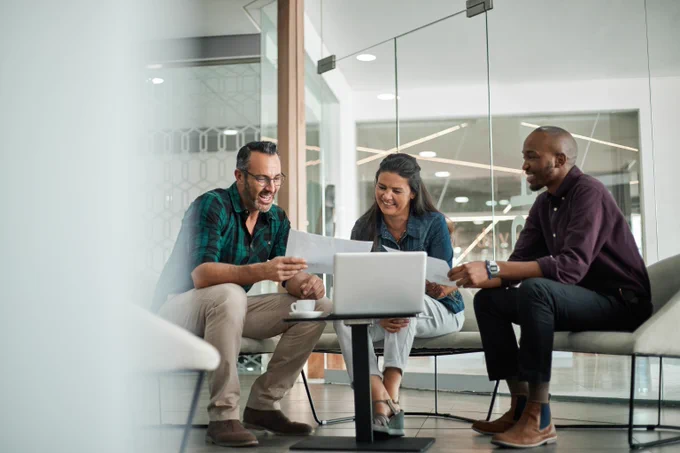 Three colleagues discussing documents around a laptop in a modern office meeting area with glass walls.