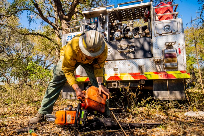 Firefighter in protective gear refills a chainsaw with fuel beside a fire truck in a wooded area.