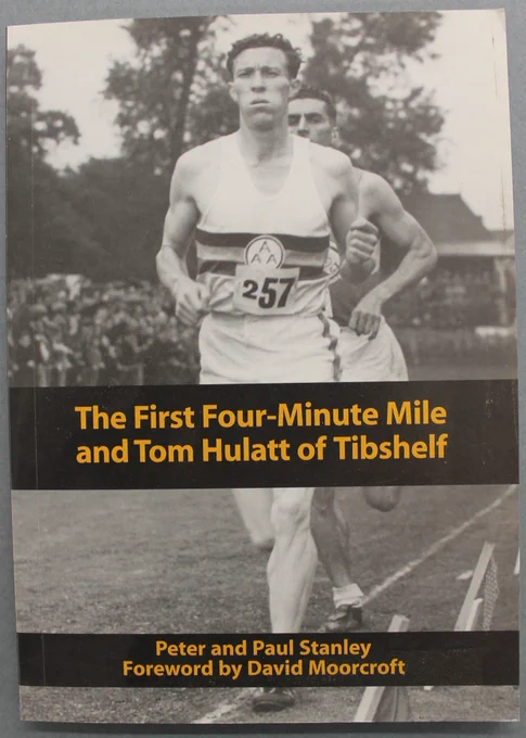 Cover of the book ‘The First Four Minute Mile and Tom Hulatt of Tibshelf’ by Peter and Paul Stanley, with a Foreword by David Moorcroft. It features a black and white photograph of a man wearing a running vest and shorts, running along a grass track.