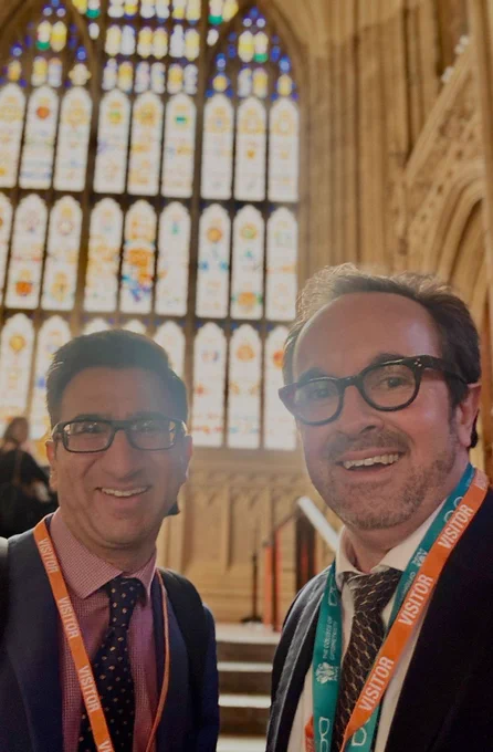 College of Optometrists staff Neal Suchak and Daniel Hardiman-McCartney MBE FCOptom smiling at an event inside the Houses of Parliment with ornate stained glass windows in the background.
