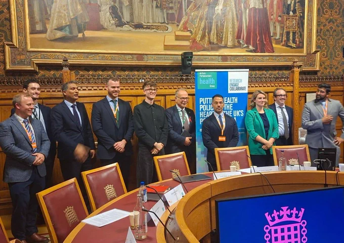 Group of individuals standing in a meeting room with a mural in the background, featuring a banner for the All-Party Parliamentary Health Group (APHG) and a digital screen with the UK Parliament symbol.