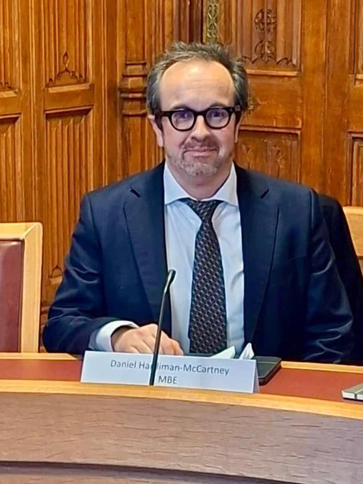 Daniel Hardiman-McCartney MBE FCOptom sitting at a desk with a nameplate reading "Daniel Hardiman-McCartney MBE", dressed in a suit, in a room with wood paneling.
