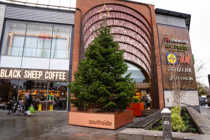 A large Christmas tree positioned in front of the Southside Shopping Centre, flanked by stores including Black Sheep Coffee with signs for Cineworld, Waitrose, and H&M.