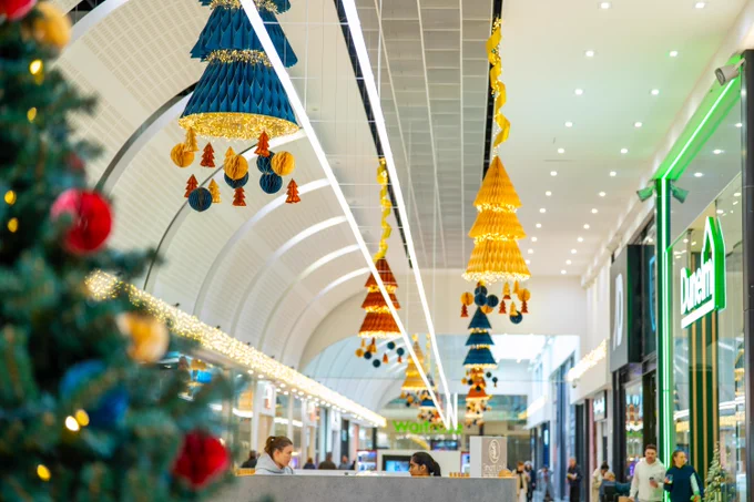 Decorated indoor shopping mall during the holiday season, featuring colourful hanging ornaments and festive lights, with shoppers visible in the background.