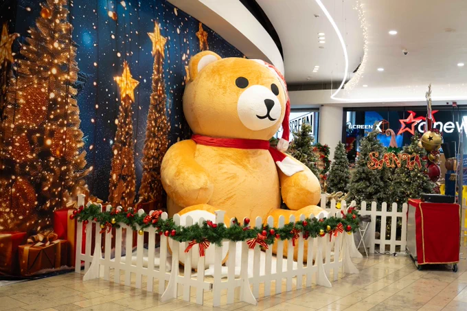Giant teddy bear with a red scarf seated beside Christmas trees inside a shopping mall, surrounded by festive decorations and a backdrop of stars.