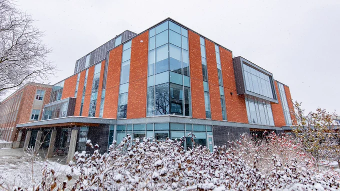 A modern building with large glass windows and orange panels, covered in light snow. There is a snow-covered bush in the foreground.