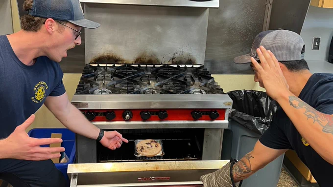 Two Austin firefighters wearing baseball caps and dark blue t-shirts peering into half-open oven door at cookie.