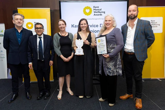 A group of six people at the awards ceremony. People are smiling and two are holding a certificate and a trophy in front of the sponsors banners