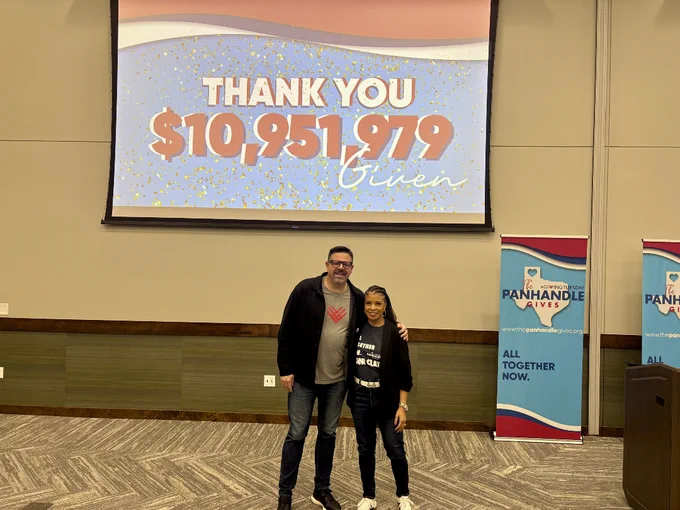 Two people stand smiling in front of a screen displaying "Thank You $10,951,979 Given." They are in a conference room with Panhandle Gives banners.