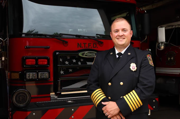 A person in a formal firefighter uniform smiling in front of a Nimishillen Township Fire Department fire truck.