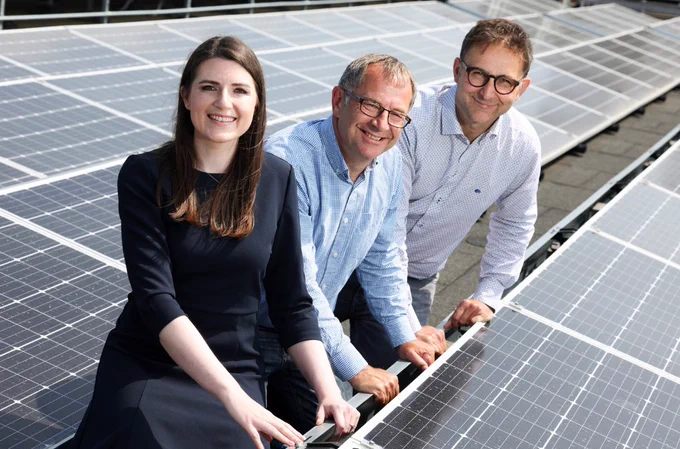 Three people, two men and a woman, are smiling and posing among a field of solar panels. The woman is wearing a black dress, and the men are in light-colored shirts. The panels reflect the sunlight, highlighting a focus on renewable energy.