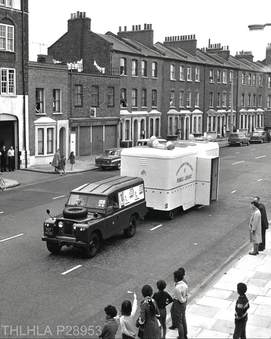 Black and white photograph of a car pulling a Mobile Library Service van while a group of children and several adults look on, taken in June 1967 on Burdett Road (ref. P28953).