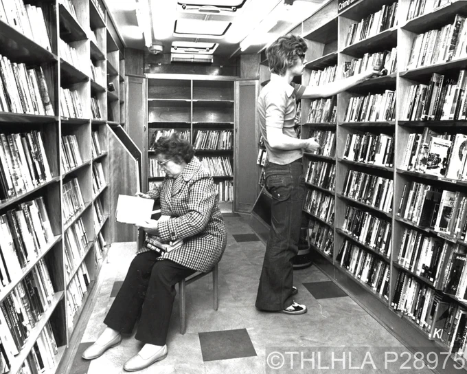 Black and white photo showing the interior of the Tower Hamlets Mobile Library Service van with a man and woman browsing the shelves, taken 20 July 1977 (© Tower Hamlets Architect's Department, ref. P28975).