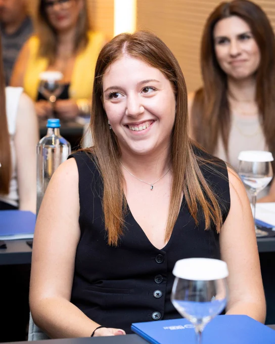 A young woman with straight light-brown hair, wearing a black sleeveless top, smiles while seated in a classroom setting with other attendees in the background.