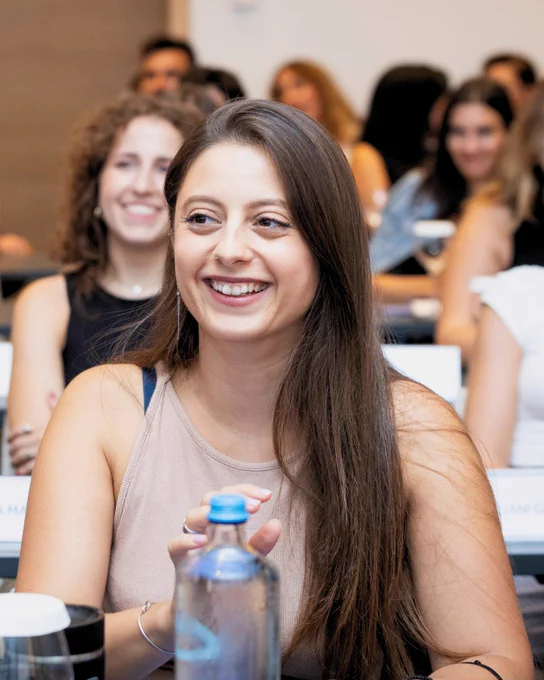 A young woman with long brown hair, smiling while seated in a classroom setting, with other attendees blurred in the background.