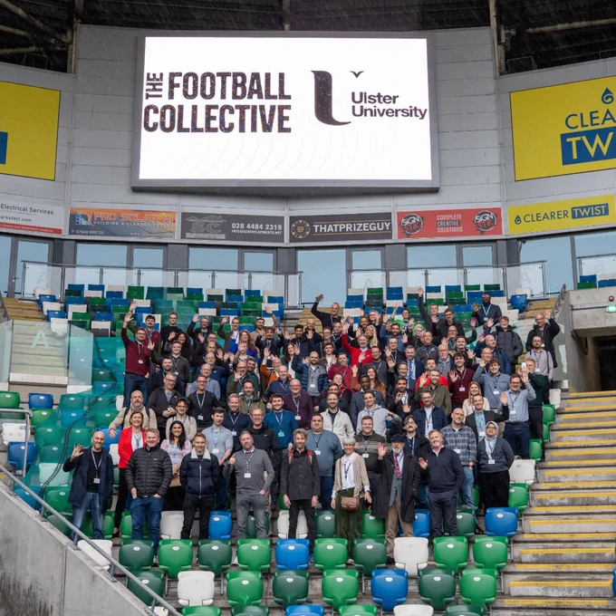 Large group over 170 delegates from over 60 institutions and 17 countries, posing in stadium seats under a screen displaying The Football Collective and Ulster University logos.