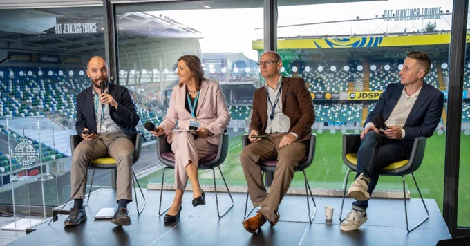 Four professionals seated on stage in front of a stadium window, engaged in a panel discussion with microphones and notes.