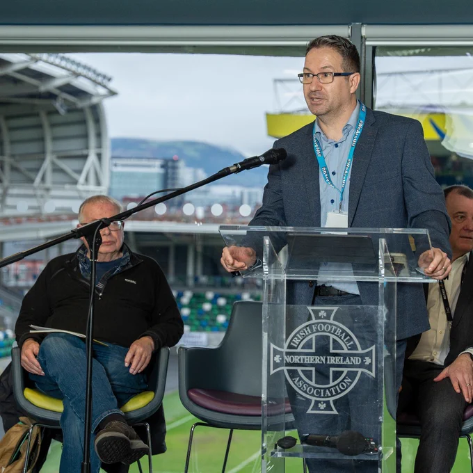 Dr Kyle Ferguson from Ulster University speaking at a clear podium with Northern Ireland Football Association logo inside a stadium lounge area.