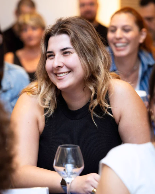 A young woman with wavy blonde hair wearing a black sleeveless top, smiling warmly while seated at a table during an event, with other attendees blurred in the background.