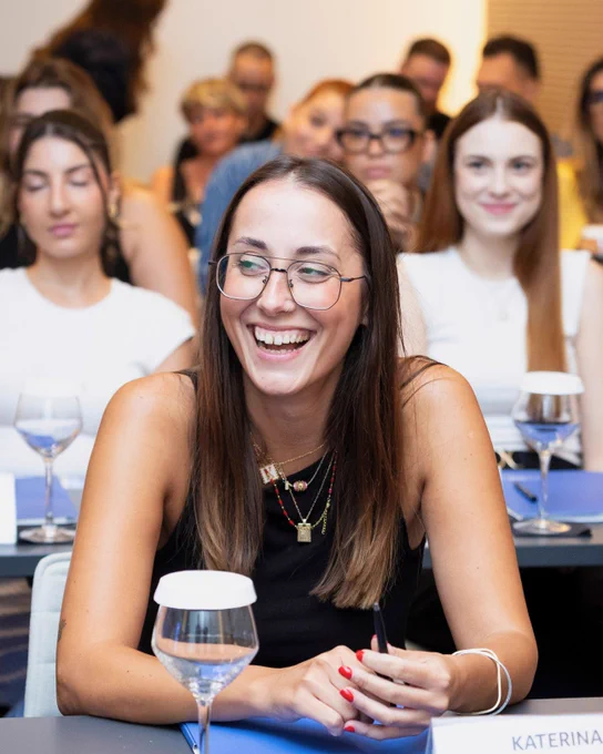 A young woman with long brown hair and glasses smiling brightly while seated at a classroom-style event, surrounded by other attendees in the background.