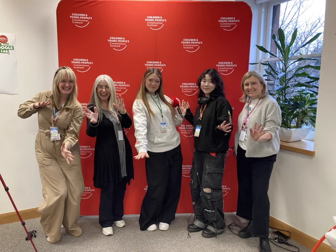 Commissioner Nicola, Dame Evelyn Glennie, Young Advisors, and Prof Laura Lundy shake in front of a red screen ahead of the Shoogle Lab.