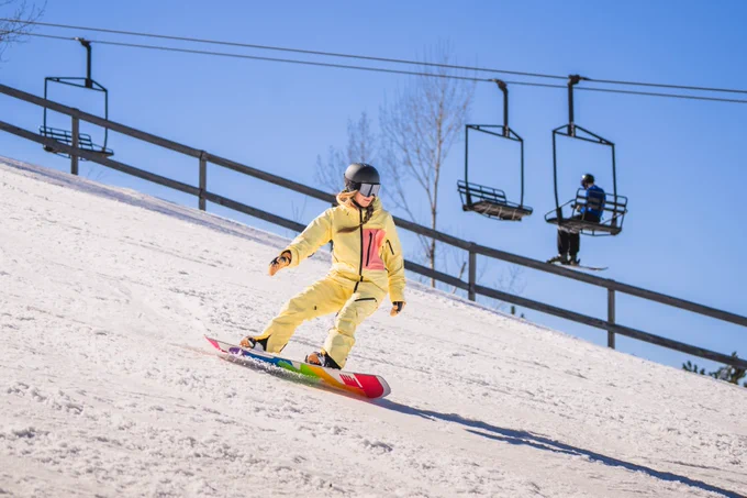 A person snowboarding down a snowy slope with chairlifts in the background under a clear blue sky.