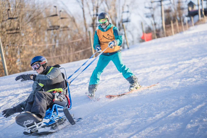 Two individuals participating in adaptive skiing on a snowy hill, with one sitting in a specialized ski sled and the other standing on skis, assisting by holding tethers attached to the sled.