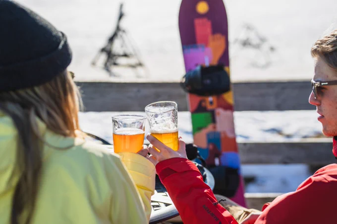Two people enjoying a beer outdoors with a snowboard in the background.