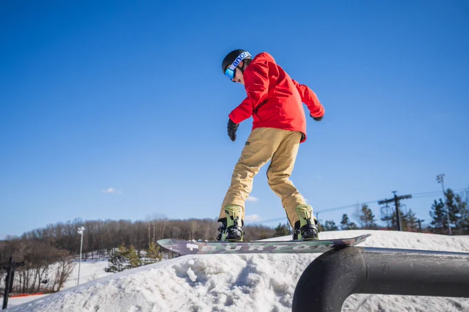 A person snowboarding, performing a trick on a rail at a snowy resort, dressed in a red jacket and tan pants. The snowboard is branded with "Burton."
