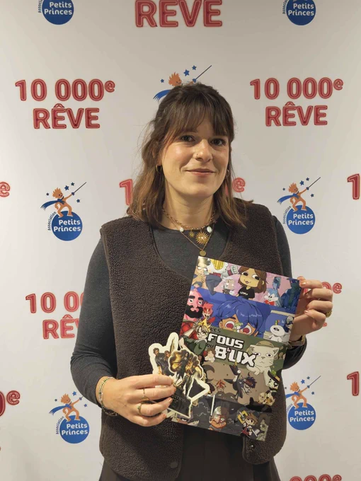 La streameuse WTF'UX pose en souriant devant le mur "10 000ème rêve" de l'Association Petits Princes avec son cadeau pour le marathon