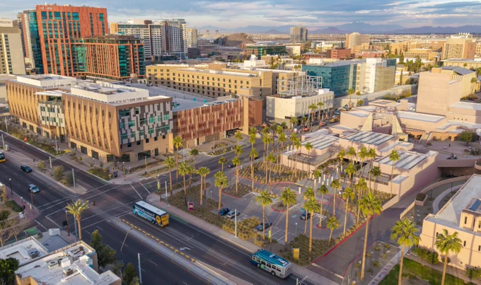 Aerial view of a cityscape featuring modern buildings, palm trees, and vehicles on a wide street under a partly cloudy sky.