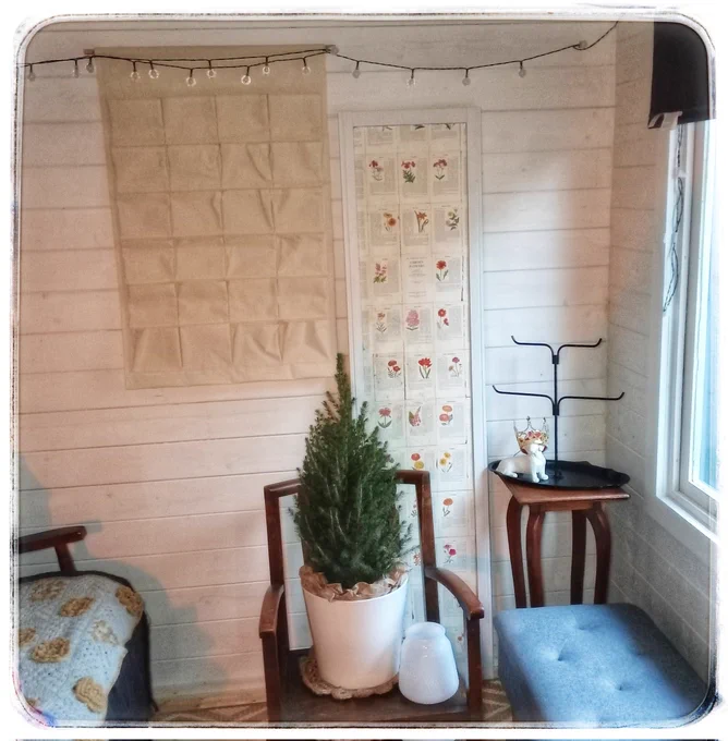 A corner of a white painted wooden shed, with solar lights strung along the top, a vintage floral book page decorated panel leaning against the wall, a plain canvas pocket advent calendar hanging, a little live tree in a white pot on an old wooden chair, & an empty black metal bauble tree on an old wooden table, with sofa & ottoman sneaking into the picture too.