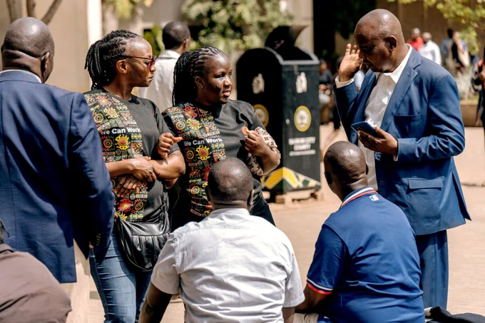 A group of adults gather in conversation during an International Day of Persons with Disabilities street awareness event in Nairobi. Two women wearing black “We Can Work” T-shirts with colorful patterned designs stand facing three men who are seated. Another man in a blue suit stands nearby, holding a phone and gesturing with his hand. The group appears engaged in a discussion along a busy urban walkway with trees, pedestrians and public bins visible in the background.