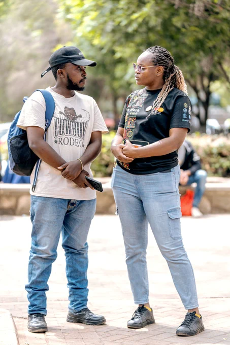 Two adults stand facing each other in conversation during a street awareness activity in Nairobi. One wears a beige T-shirt with a cowboy graphic, a black cap and a backpack, holding a phone in both hands. The other wears a black “We Can Work” T-shirt with colorful patterns and light blue jeans, speaking attentively. They stand in a shaded outdoor area with trees and people in the background.