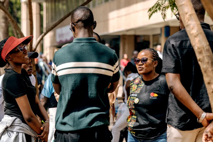 A small group of people stand together talking during an International Day of Persons with Disabilities street awareness event in Nairobi. One woman wearing a black “We Can Work” T-shirt with colorful patterns and sunglasses speaks while facing three men. Another woman in a red visor and black shirt listens with her hands clasped. The group stands under partial shade along a busy sidewalk with buildings and passersby in the background.