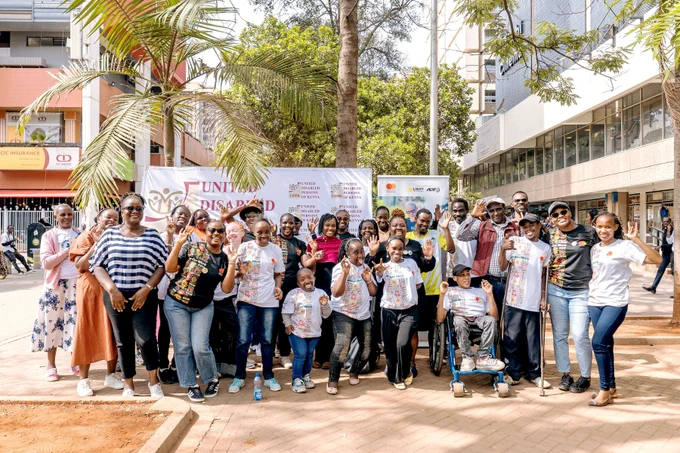 A large, cheerful group of participants pose for a photo during the International Day of Persons with Disabilities street awareness event in Nairobi. Adults and youth including wheelchair users and people using mobility aids, stand together in front of a United Disabled Persons of Kenya banner. Many wear “We Can Work” T-shirts with colorful patterns and raise their hands while smiling at the camera. The scene is set on a lively city street with buildings, trees and passersby in the background.