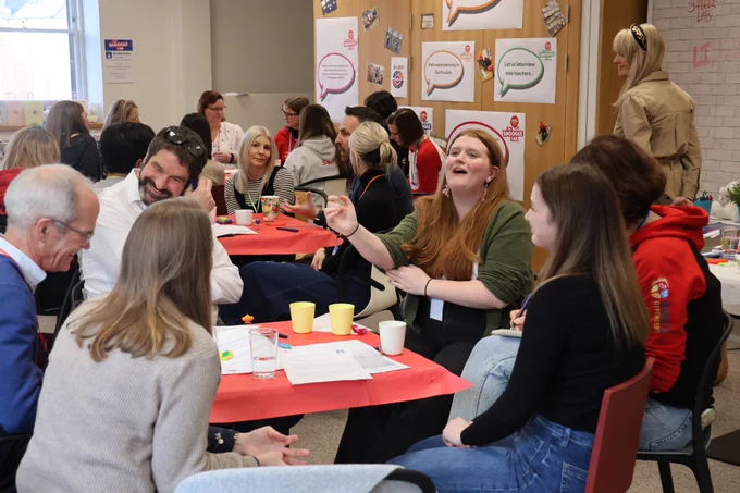 A group of adults sit around tables covered with red paper, talking and laughing during a workshop. One woman in a green top is speaking animatedly, while others listen and smile. Papers, cups, and markers are spread across the tables. Posters about children’s voices and education hang on the wall in the background.