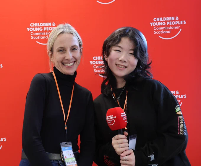 Two people stand smiling in front of a bright red backdrop with the Children and Young People’s Commissioner Scotland logo. On the left is an adult woman with light hair tied back, wearing a black top and an orange lanyard. On the right is a young person with dark wavy hair, holding a red CYPCS-branded microphone and wearing a black top with a lanyard.