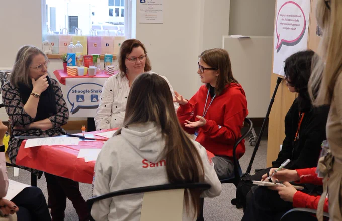 A young advisor in a red hoodie speaks while gesturing with both hands during a group discussion. Several adults and young people sit in a circle listening, taking notes, and engaging. The table is covered in red paper with worksheets on top, and a refreshment table with drinks is visible in the background. A sign reading “Shoogle Shaker” is partly visible behind the group.