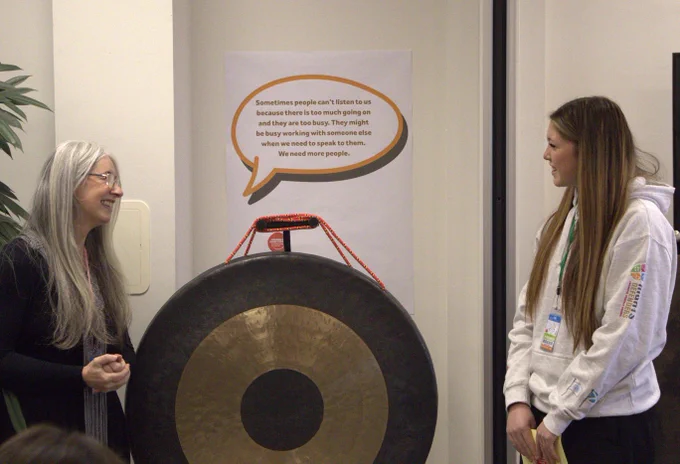 A young person in a light grey hoodie stands smiling beside a large brass gong, facing an adult woman with long grey hair who is also smiling. Behind them is a speech-bubble poster featuring a child’s quote about adults being too busy to listen.