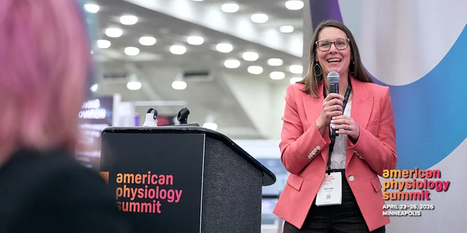 Speaker in a coral blazer holding a microphone at the American Physiology Summit podium.