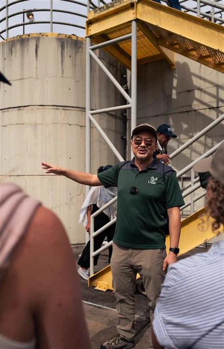 Person in green polo shirt gesturing while speaking to a group near industrial stairs and large storage tanks.