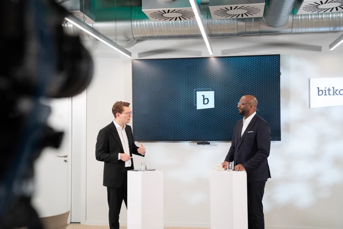 Links im Bild ist Fabian Zacharias, Mitglied der Geschäftsführung bei Bitkom, rechts steht Armand Zorn, der stellvertretende Fraktionsvorsitzenden der SPD-Bundestagsfraktion. Die beiden Männer in dunklen Anzügen stehen sich in einem hellen Konferenzraum gegenüber und führen ein Gespräch. Beide stehen hinter weißen Stehpulten mit Wassergläsern. Im Hintergrund ist ein großer Bildschirm mit dem Logo eines stilisierten Buchstabens „b“ zu sehen. Rechts an der Wand hängt ein weiteres Schild mit dem gut lesbaren Schriftzug „bitkom“. Eine Kamera ist am linken Bildrand angeschnitten und deutet auf eine Live- oder Videoaufnahme hin. 