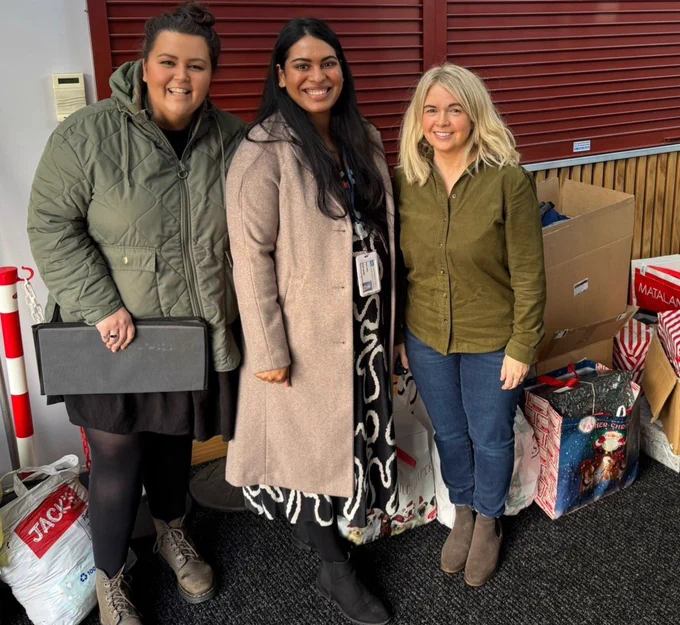 Three individuals smiling, standing together in a room surrounded by christmas gifts
