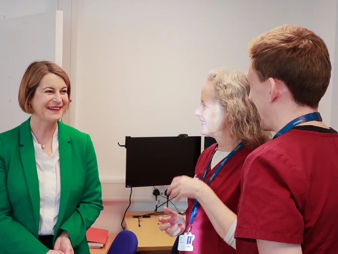 Three individuals interact cheerfully in an office setting; Helen Hayes MP wearing a bright green blazer and the other two in red scrubs with ID badges, standing near a computer desk.